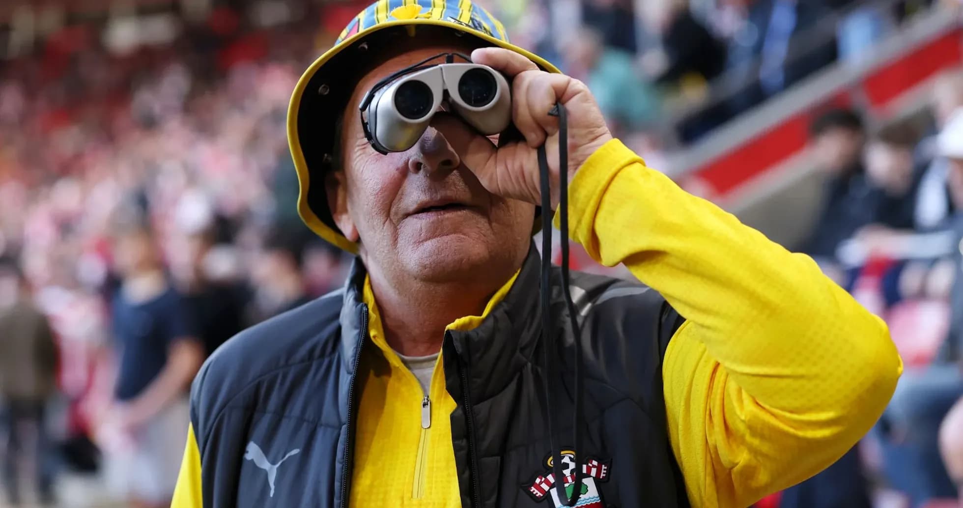 A fan wearing binoculars and a hat at a match
