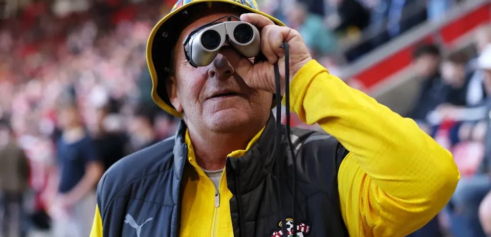 A fan wearing binoculars and a hat at a match